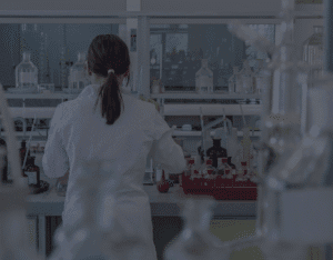 A female laboratory technician in a white lab coat working at a laboratory bench surrounded by glassware, chemical bottles, and scientific equipment.