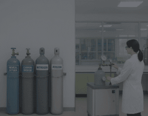 A female lab technician in a white coat adjusts a regulator on a purified gas cylinder inside a modern laboratory. Four large cylinders labeled Medical Oxygen, Carbon Dioxide, Nitrogen, and Argon are securely chained against the wall, with the banner text reading “Purified Gases: Powering Healthcare, Research, and Industry in Malaysia.