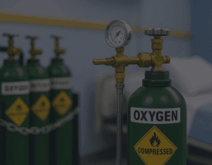 Four green medical oxygen cylinders with pressure gauges and warning labels, securely chained in a hospital room next to a patient bed, representing safe and reliable oxygen supply in Malaysia.