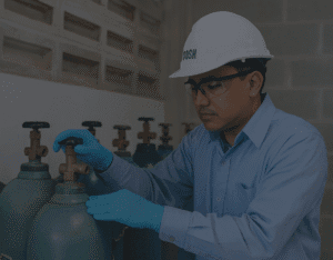 A DOSH-certified Malaysian technician wearing safety glasses and gloves inspects a gas cylinder valve in a secure, well-ventilated storage area to ensure safety compliance.